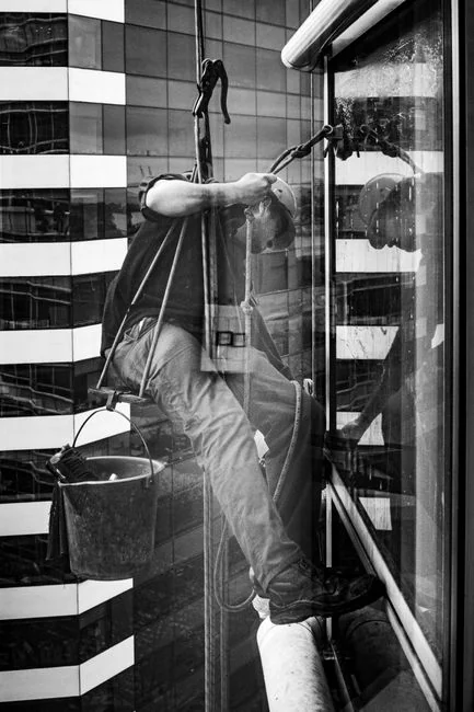 Vue en noir et blanc, prise depuis le bas, d’un homme cordiste portant un casque de protection et équipé de matériel professionnel, nettoyant la façade vitrée d’une tour moderne